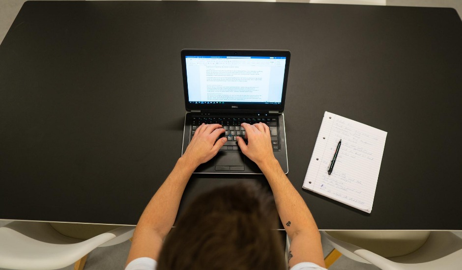 Aske skriver på computer ved et skrivebord. Der ligger en blok og en kuglepen ved siden af ham. Kjærup Kommunikation er hans arbejdsplads.